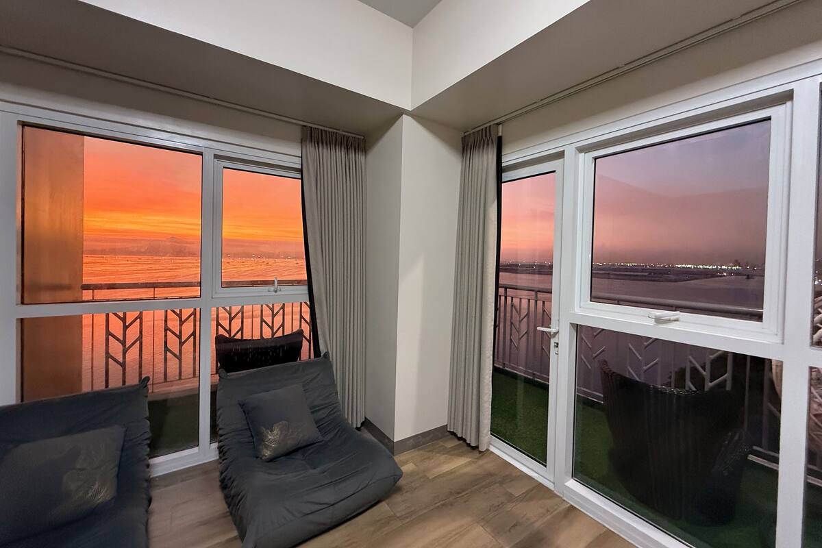 A balcony with a view of a sunset over water, a cushioned seating area, and a window with a view of a cityscape.