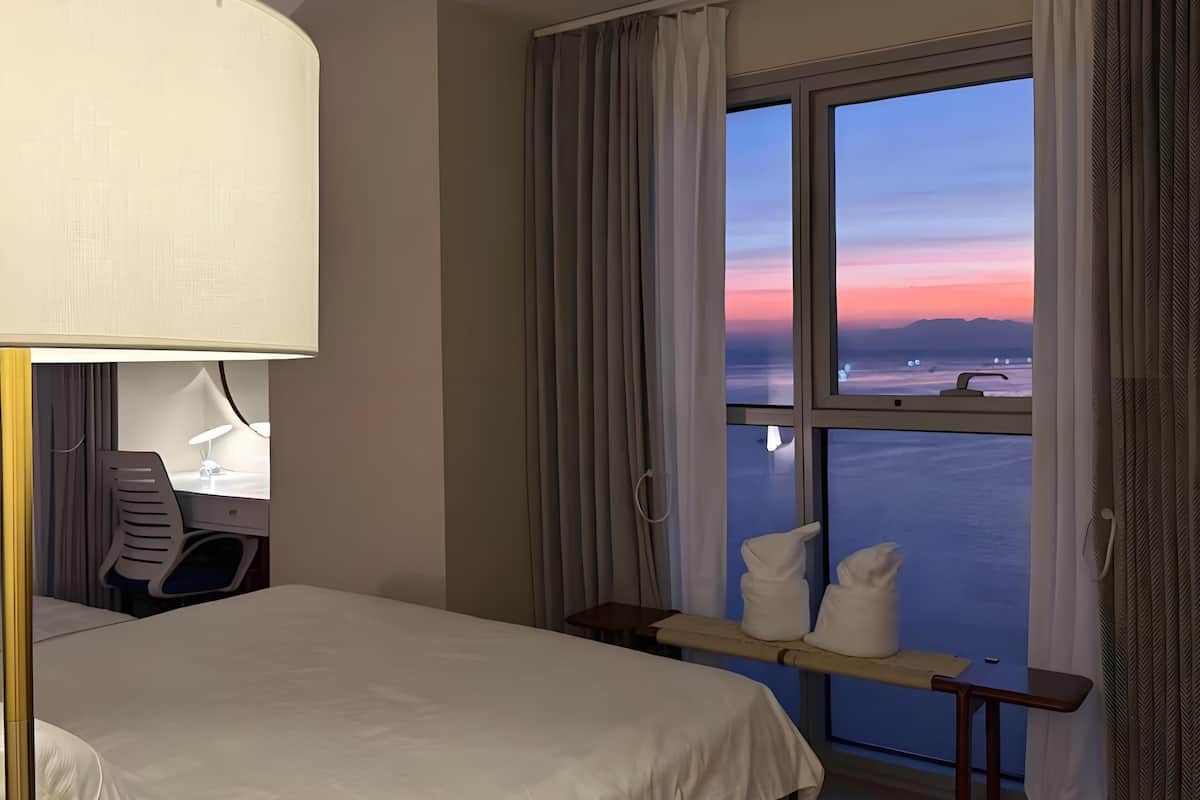 A hotel room with a bed, a desk with a chair, a lamp, and a view of the sunset over the water.