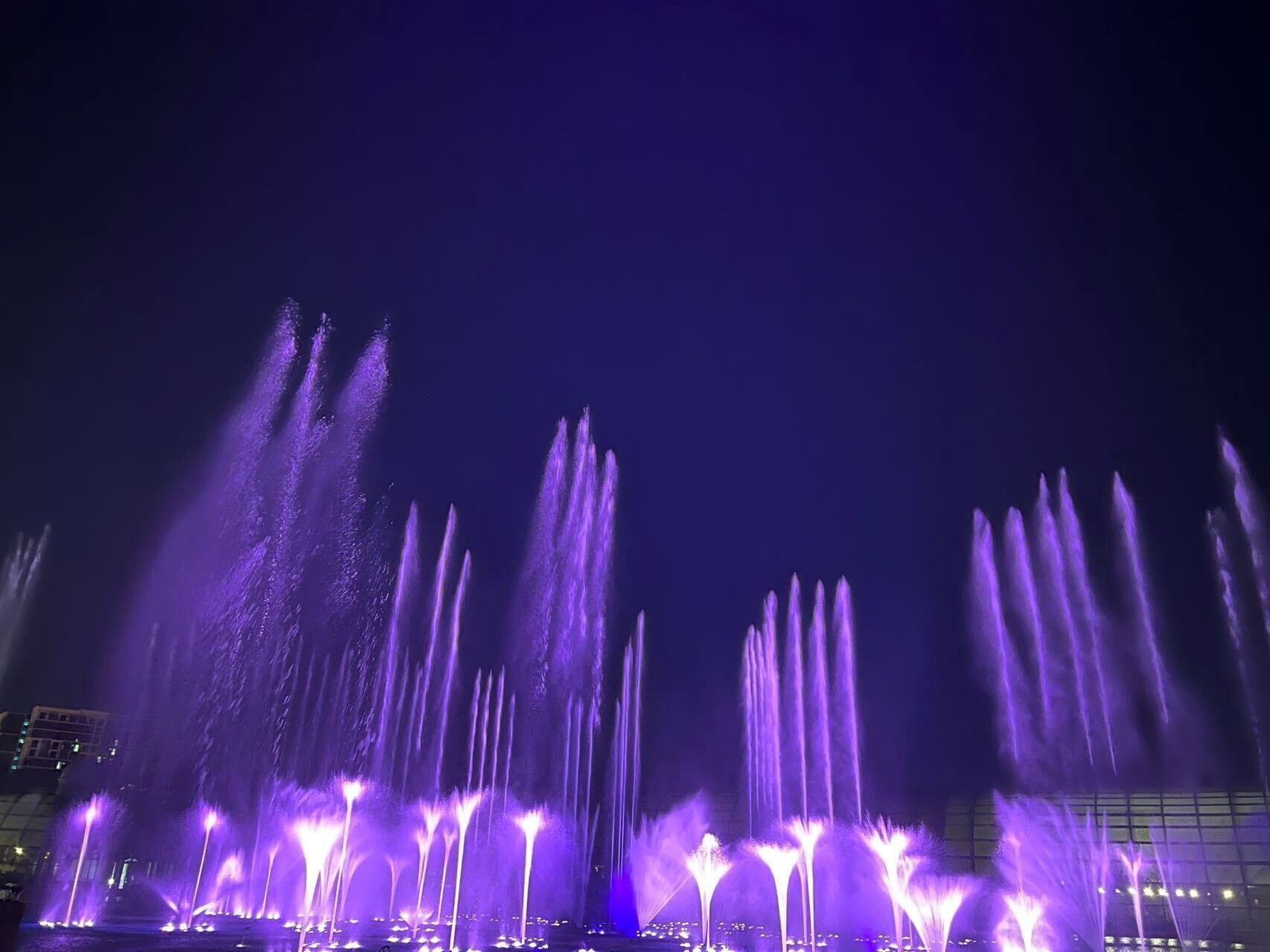 A nighttime water fountain display with multiple jets of water illuminated in purple.