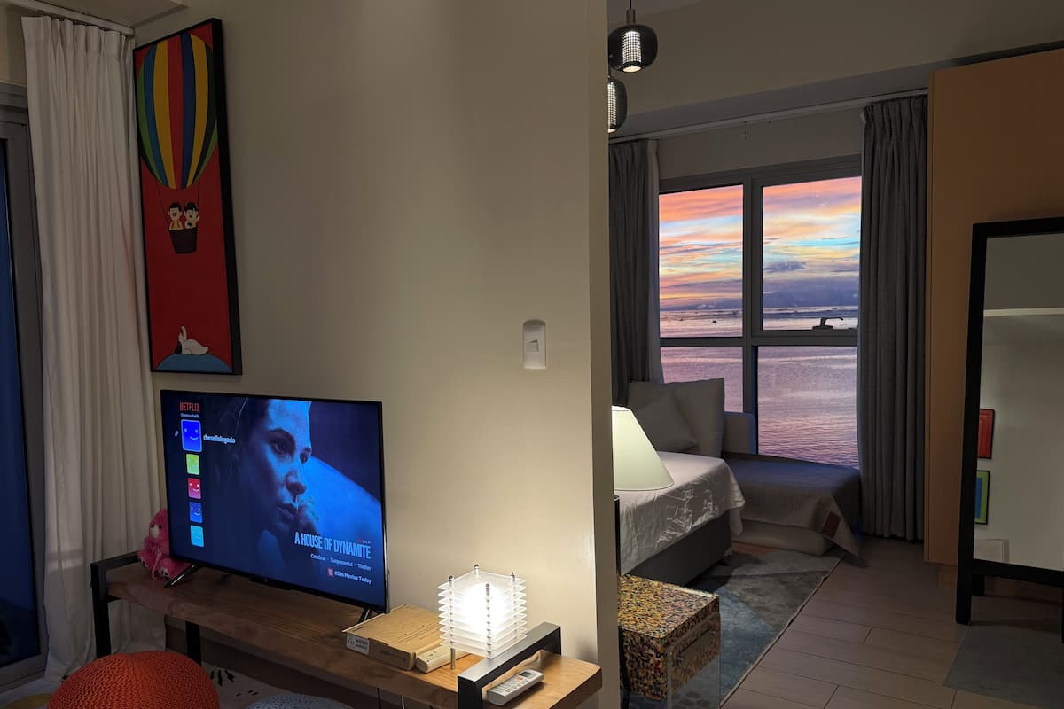 A living room with a TV, a cozy seating area, a window with a sunset view, and a small table with a lamp.