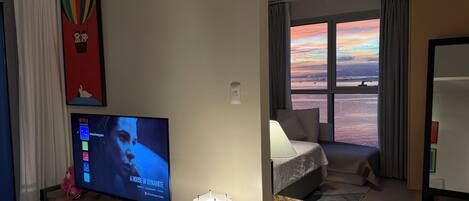 Ein Wohnzimmer mit Fernseher, einer gemütlichen Sitzecke, einem Fenster mit Sonnenuntergangsblick und einem kleinen Tisch mit Lampe.