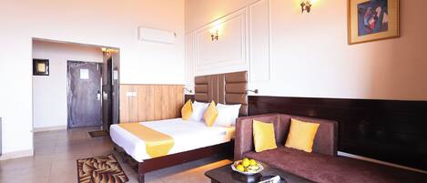 A hotel room with two beds, a sofa, a coffee table with fruits, and a painting on the wall.