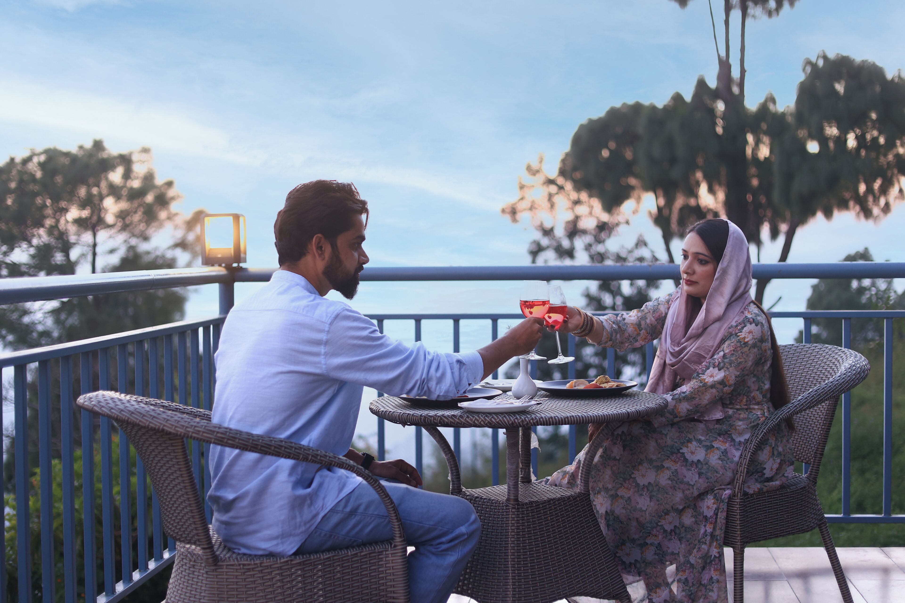 Two people dining at an outdoor table with wicker chairs, overlooking a body of water and trees.