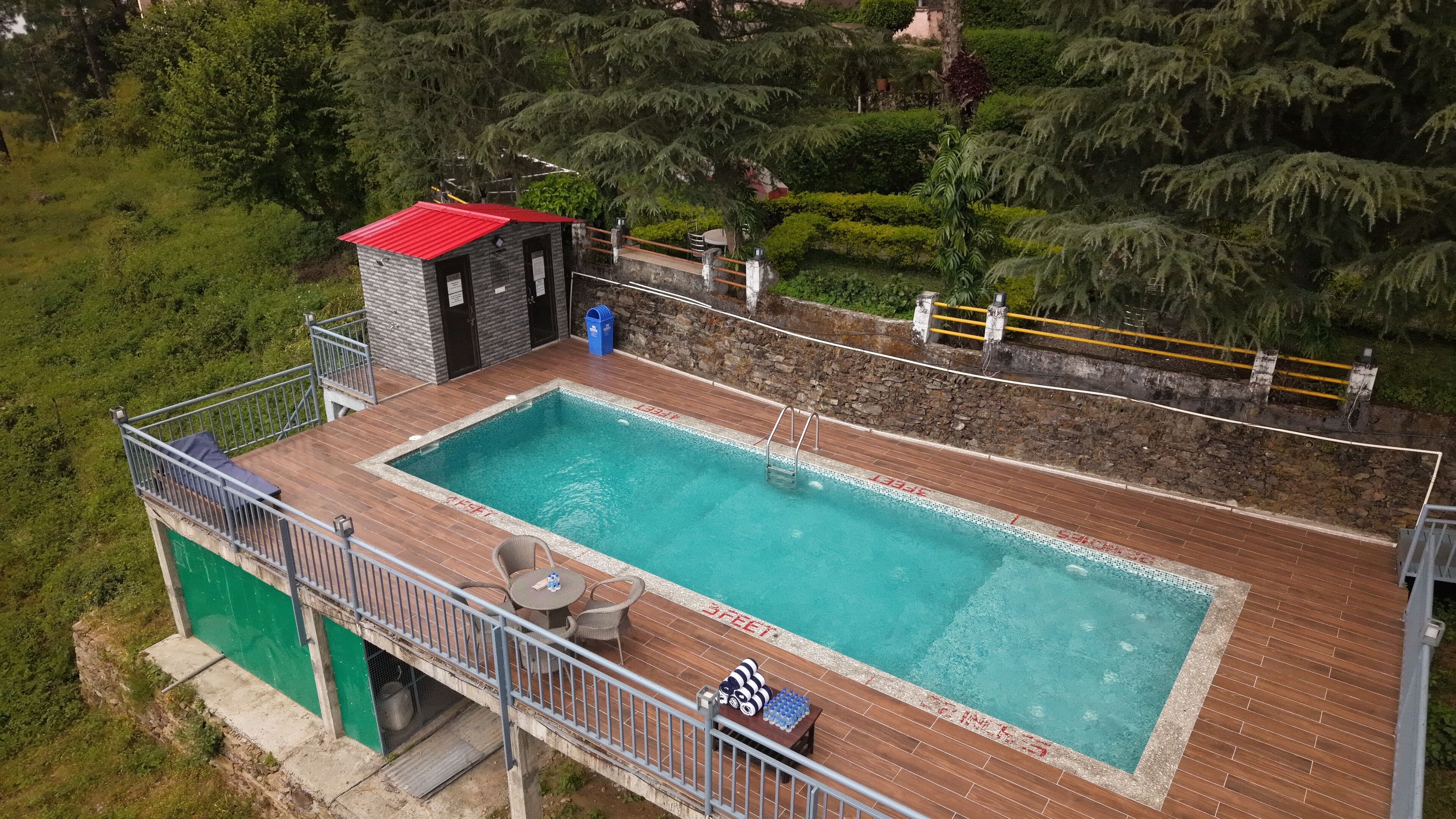 Une piscine rectangulaire avec une terrasse en bois, un petit bâtiment doté d’un toit rouge et un mur en pierre.