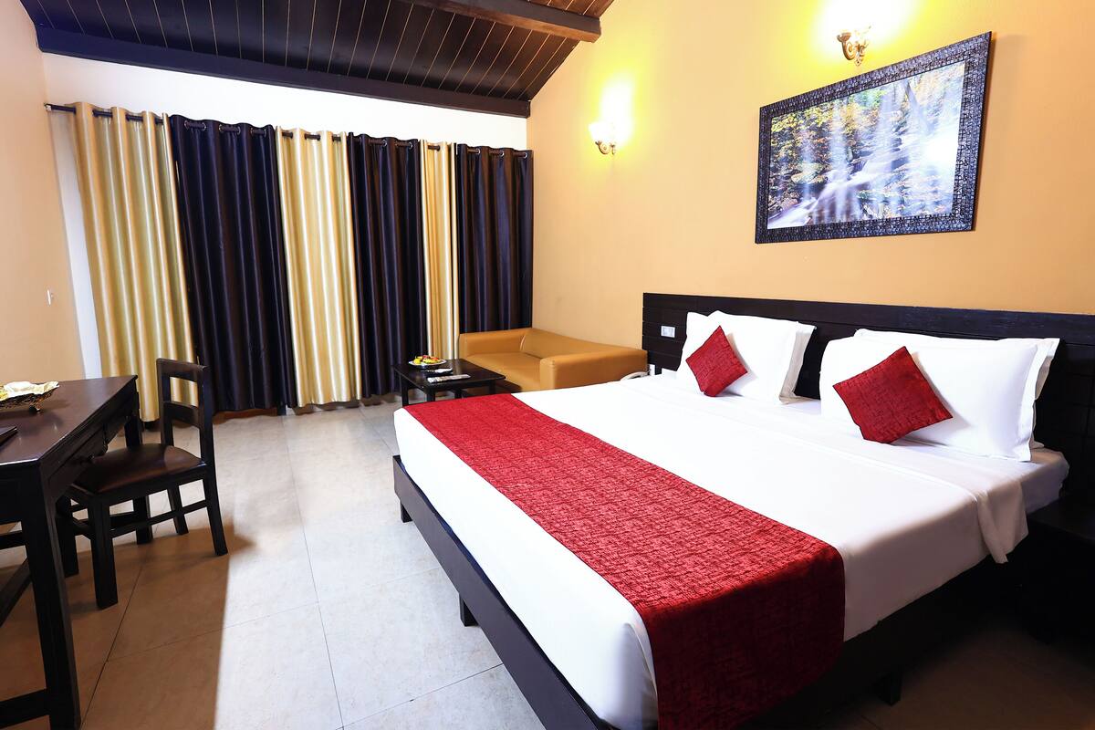 A hotel room with a large bed, a desk, and a chair. There is a painting on the wall and a window with curtains.