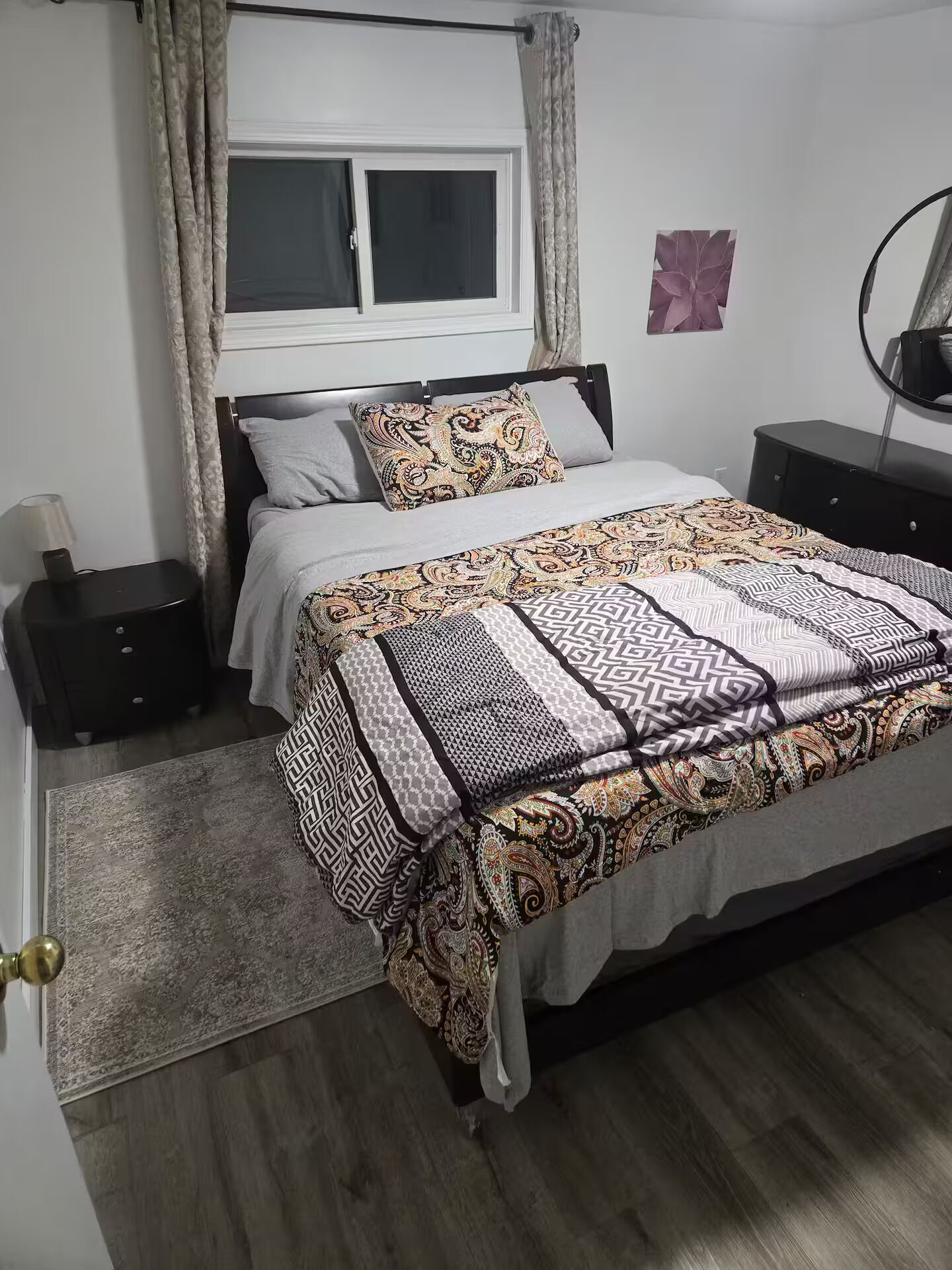 Une chambre à coucher comprenant un lit, une table de chevet, une commode, une fenêtre avec des rideaux et un miroir fixé au mur.