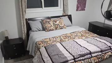 Ein Schlafzimmer mit einem Bett, einem Nachttisch, einer Kommode, einem Fenster mit Vorhängen und einem an der Wand befestigten Spiegel.