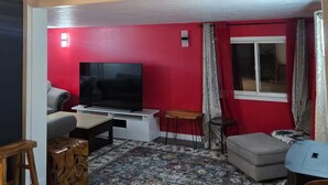 Een woonkamer met een rode muur, een flatscreen televisie, een grijze bank, een houten salontafel en een kamerbreed tapijt met een patroon.