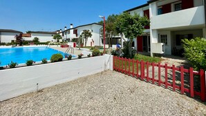 A swimming pool surrounded by a red fence, with white buildings and a clear blue sky in the background. - Terraced House With Garden and Pool (Caorle)