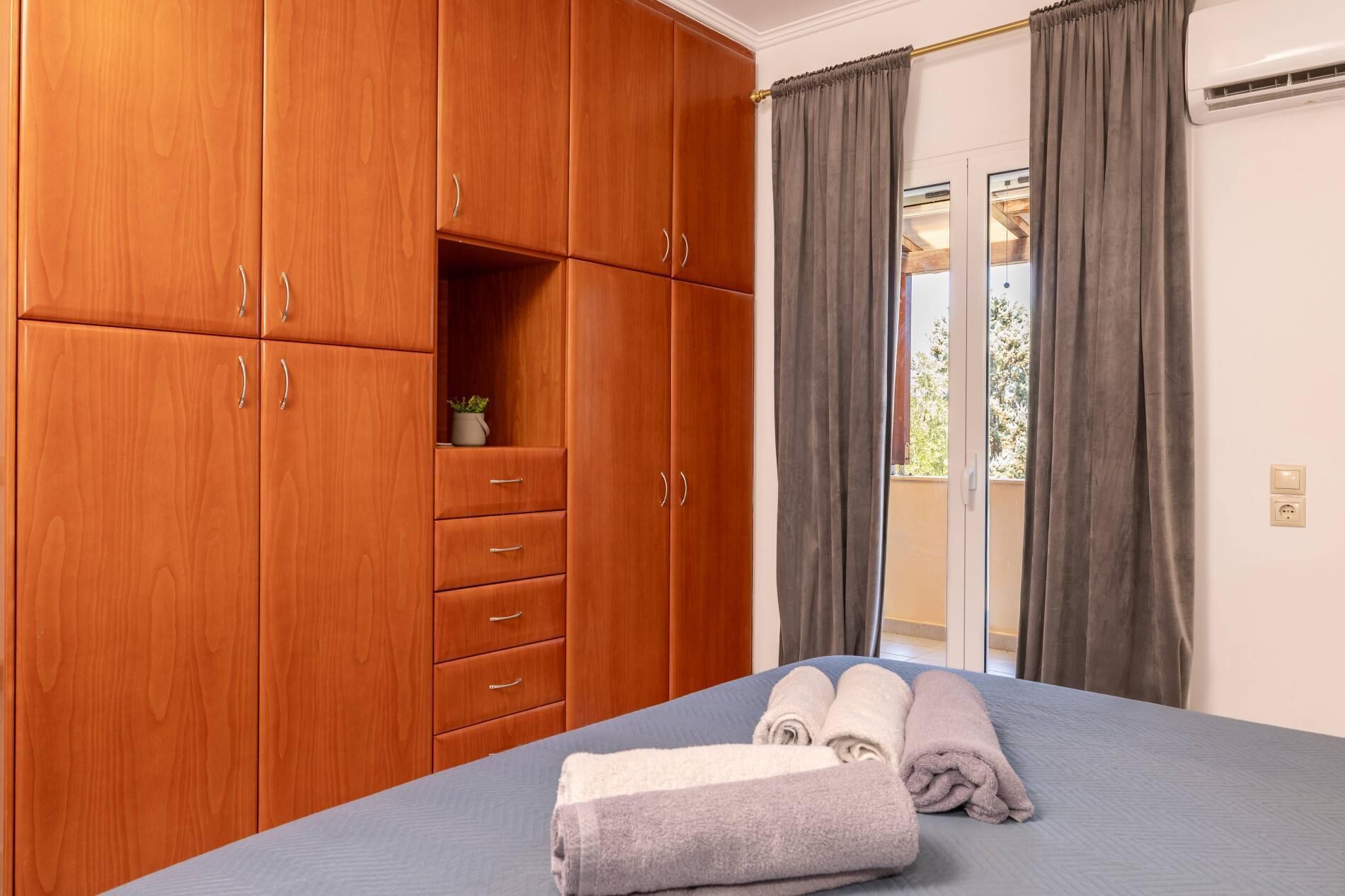 Una camera da letto con un armadio in legno, un letto con asciugamani arrotolati e una porta a vetri scorrevoli verso un balcone.