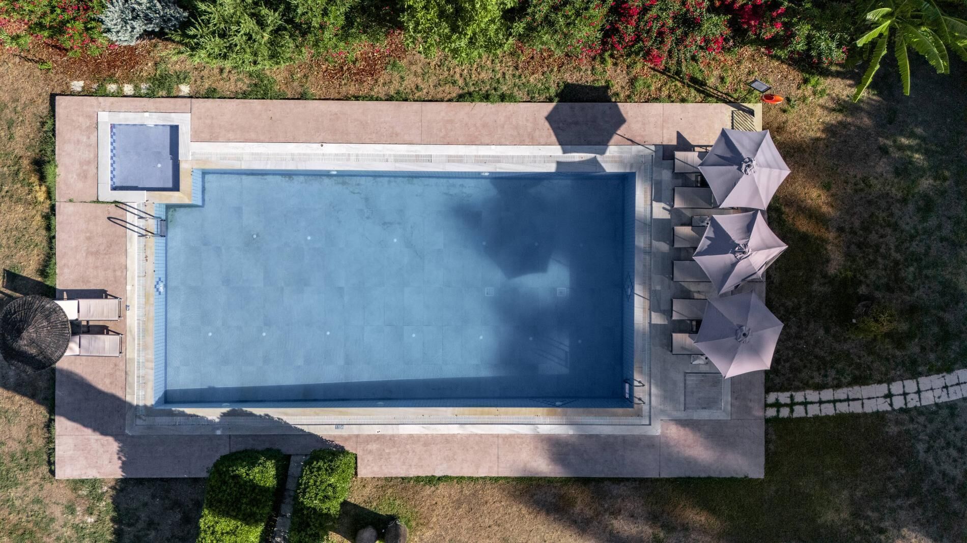 Vista aerea di una piscina rettangolare con bordo piastrellato, circondata da un basso muro in pietra. Su un lato, diversi ombrelloni e lettini prendisole, e sull’altro, una piccola zona relax con tavolo e sedie.