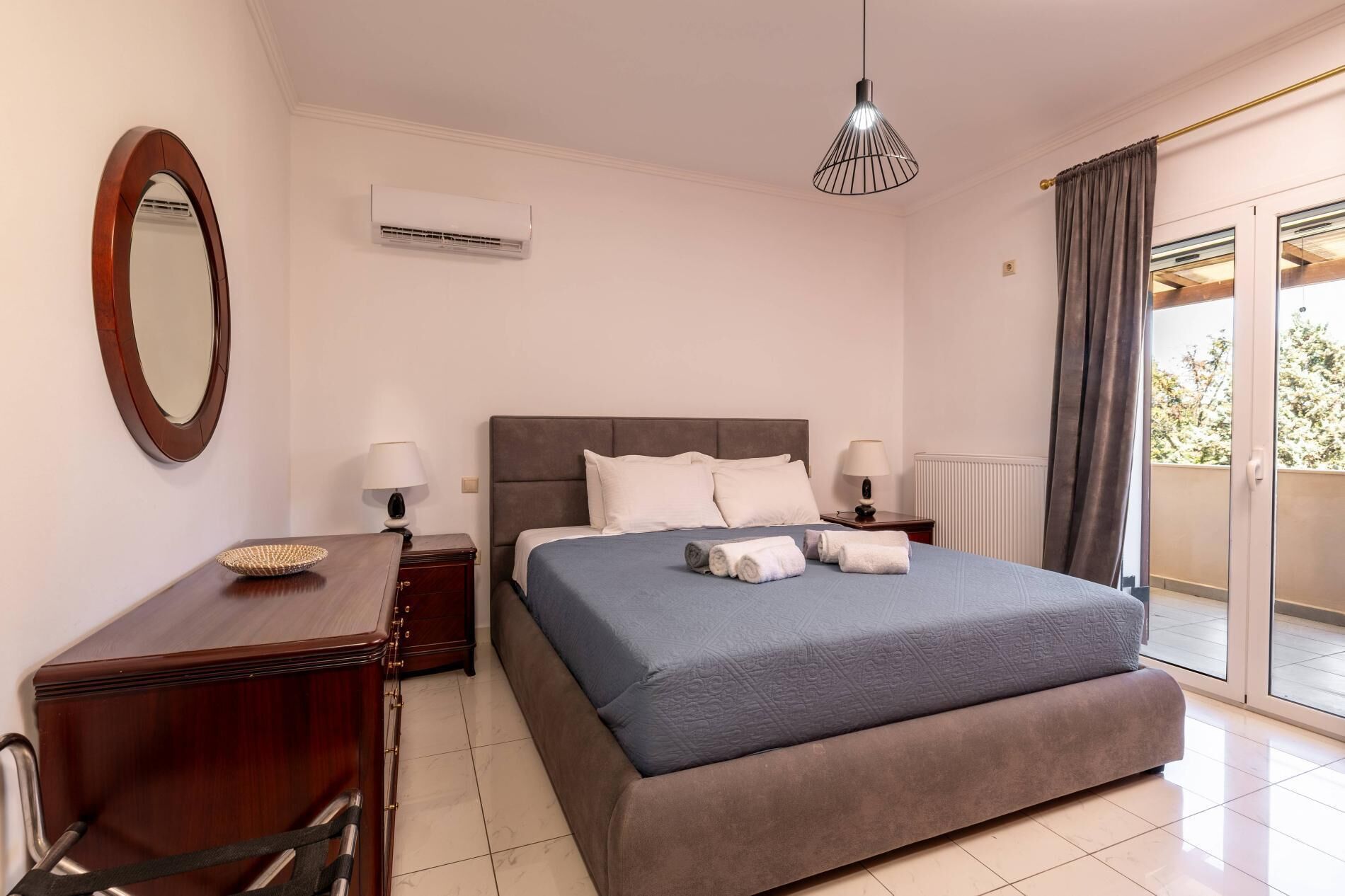 A bedroom with a large bed, two bedside tables, a mirror, an air conditioning unit, and a view of the outdoors.