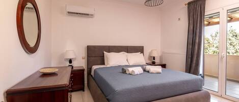A bedroom with a large bed, two bedside tables, a mirror, an air conditioning unit, and a view of the outdoors.