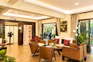 Een hotellobby met een receptiebalie, een zithoek met stoelen en banken, en een groot raam met uitzicht op groen.
