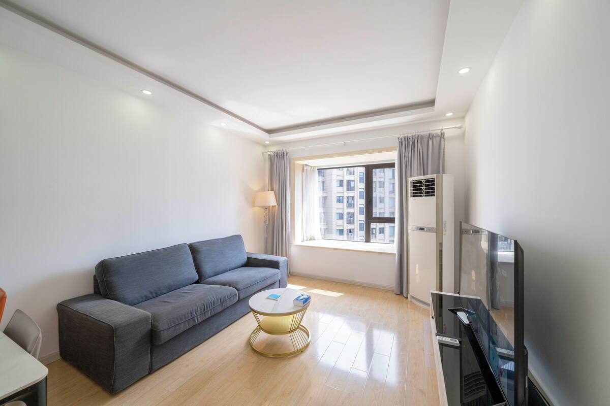 A modern living room with a grey sofa, a small round coffee table, a flat-screen TV, and a window with curtains.