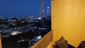 Un balcón con vistas a un paisaje urbano nocturno, destacando un edificio alto con luces. Se observan dos sillas y una mesita con una planta.