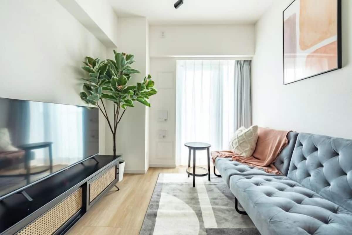 Uma sala de estar moderna com uma TV de tela plana, um sofá cinza com capitonê, uma pequena mesa redonda e uma planta grande.