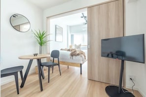 Quarto de hotel moderno com uma cama, televisão, uma mesinha com cadeiras e um vaso de planta.