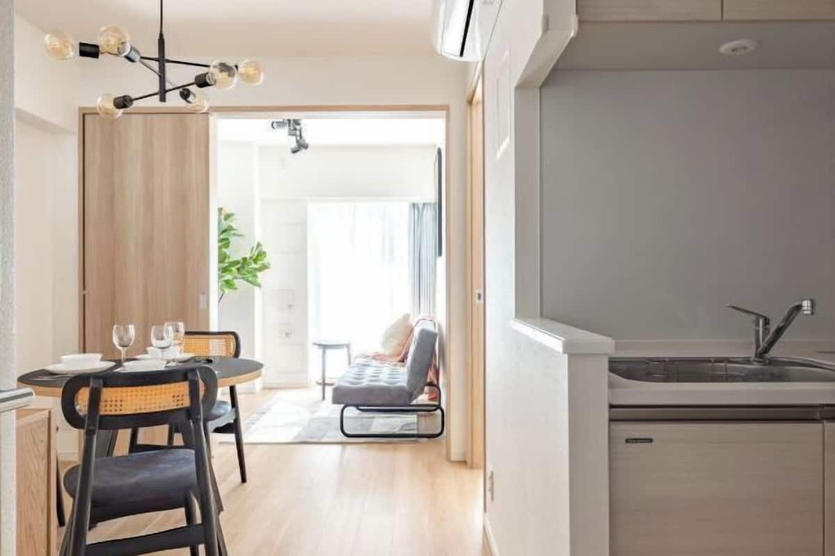 Uma cozinha moderna com máquina de lavar louça embutida, uma área de jantar com mesa redonda e cadeiras, e vista para uma sala de estar com sofá e planta.