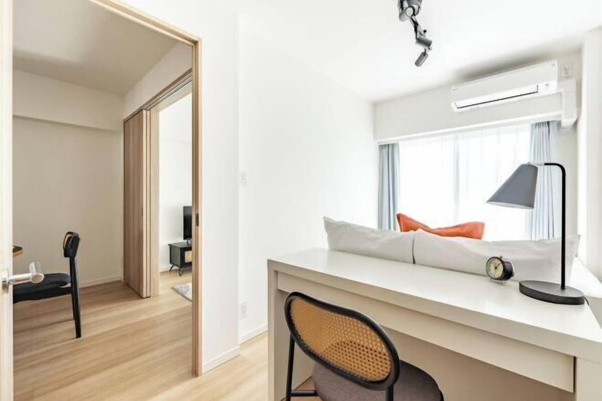 Quarto de hotel moderno com escrivaninha branca, cadeira, cama com almofadas, janela com cortinas, ar condicionado e pequena mesa com abajur.