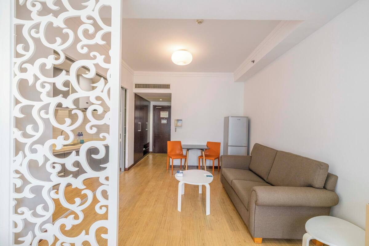 En moderne stue med en sofa, et lille bord og to stole. I baggrunden er der et køkken med et køleskab og en vask.