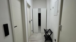 A hallway with white walls and a light wood floor, leading to a dark room at the end.