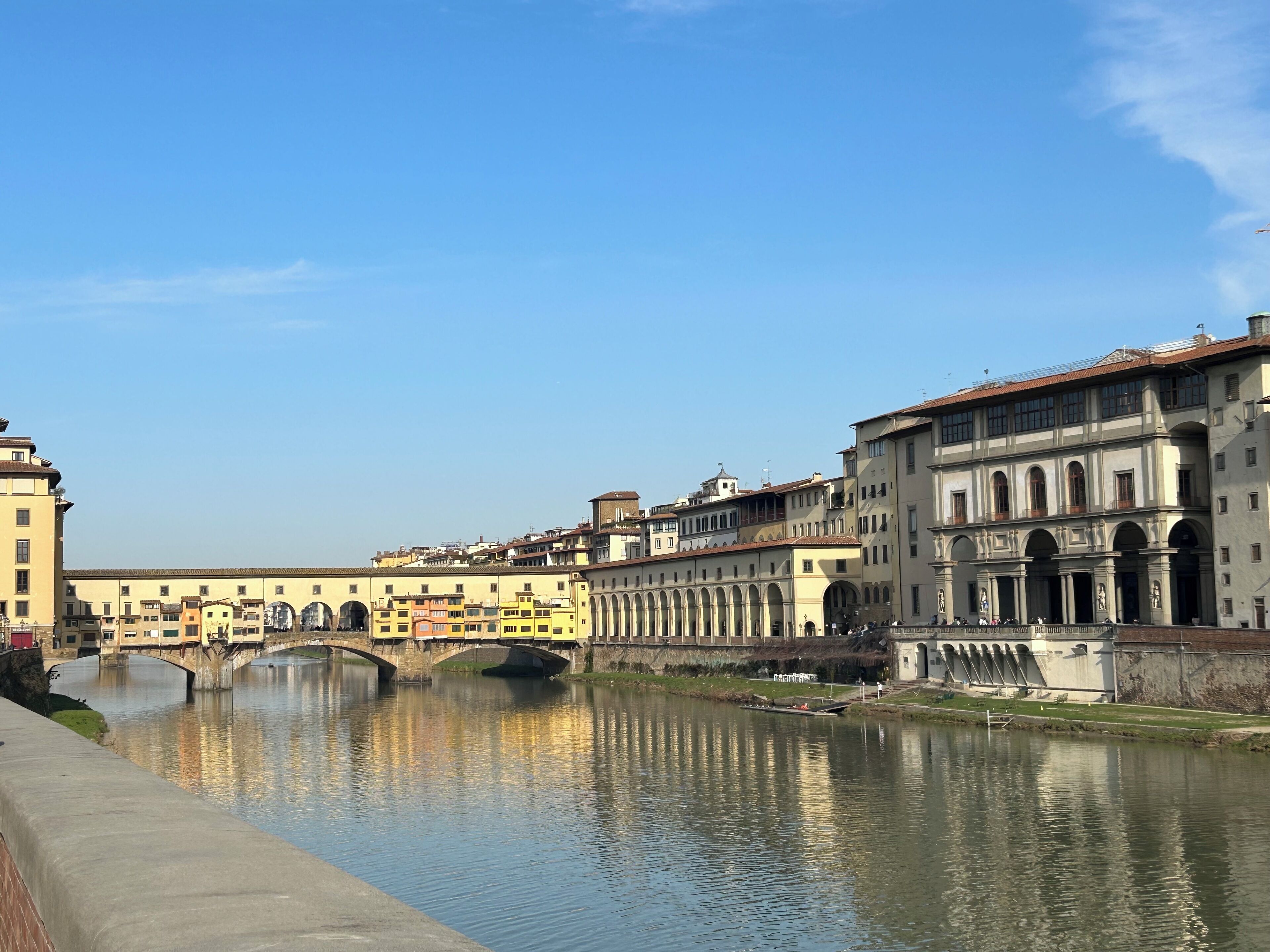 A river with a stone bridge, buildings with arches, and a clear sky.