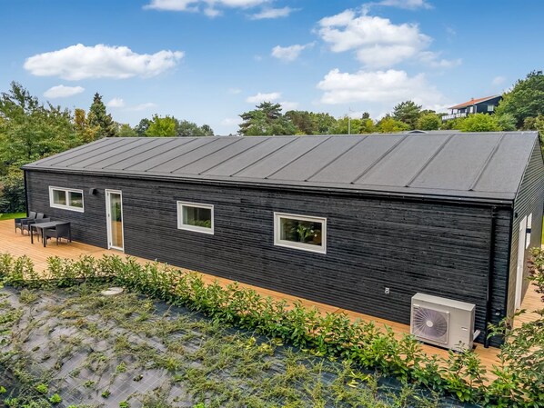 Un bâtiment moderne, de plain-pied, avec une façade sombre, un toit plat et un petit patio aménagé avec du mobilier d’extérieur.