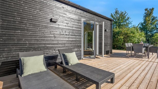 Eine Holzterrasse mit zwei Liegestühlen und einem Tisch, eine Glastür, die zu einem Außenbereich führt, und ein Gebäude mit dunkler Holzfassade.