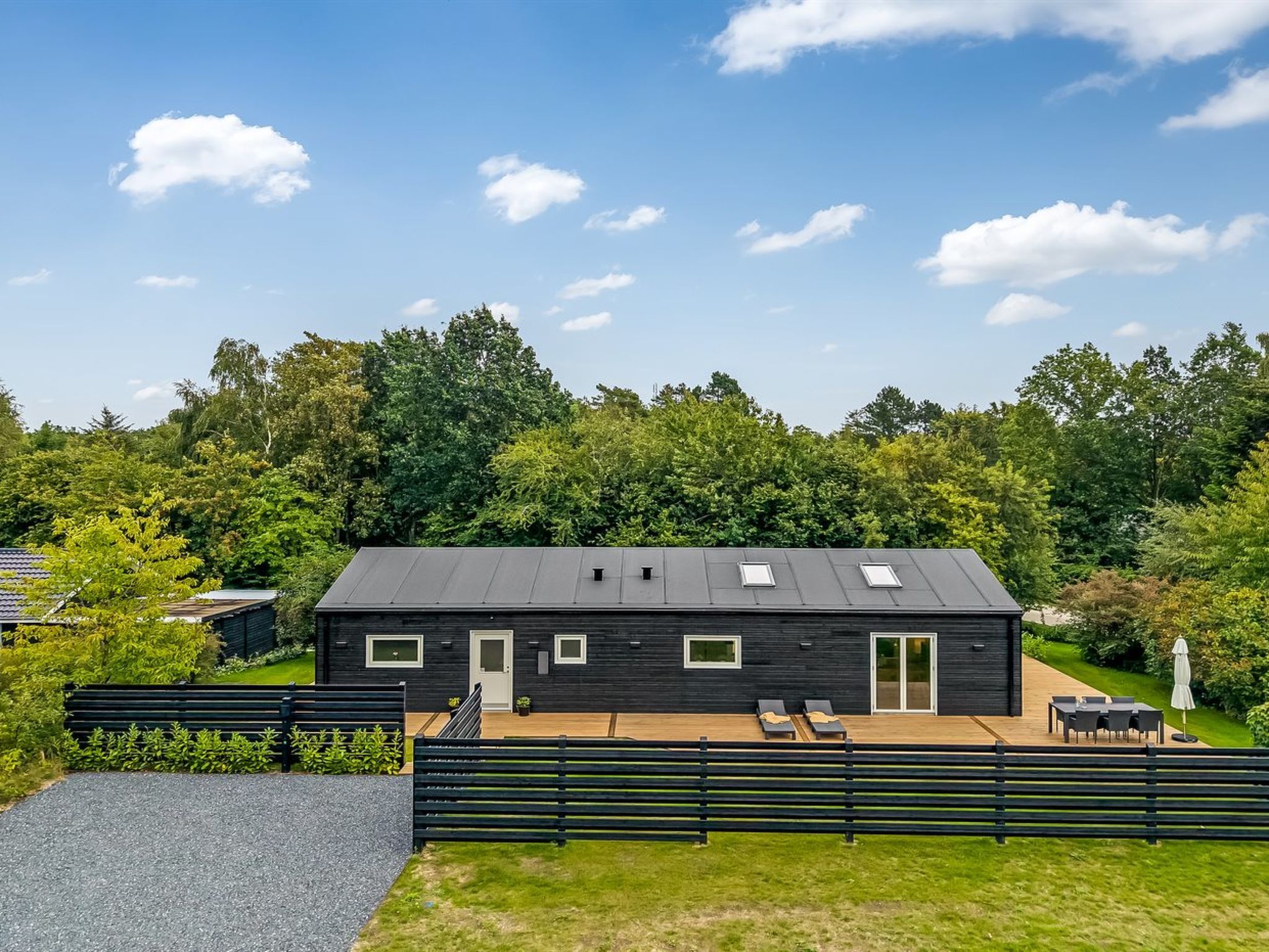 A modern black house with a flat roof, a patio with outdoor furniture, and a surrounding wooded area.