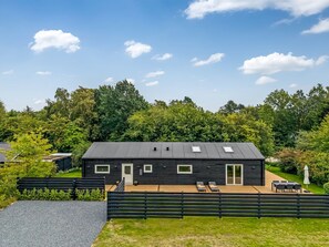 Une maison moderne à la façade noire, dotée d’un toit plat, d’une terrasse avec mobilier d’extérieur et entourée d’une zone boisée.