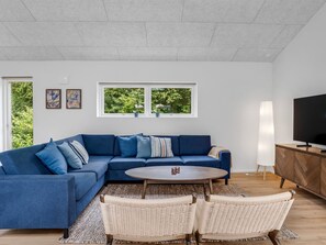 Un salon moderne avec un canapé d’angle, une table basse ronde, des fauteuils en osier, un téléviseur et une grande fenêtre donnant sur un paysage verdoyant.