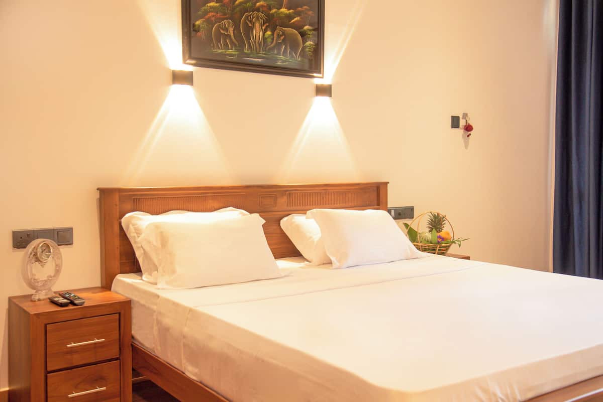 A neatly made bed with white linens and pillows, a wooden nightstand with a lamp and remote controls, and a framed picture on the wall above the bed.