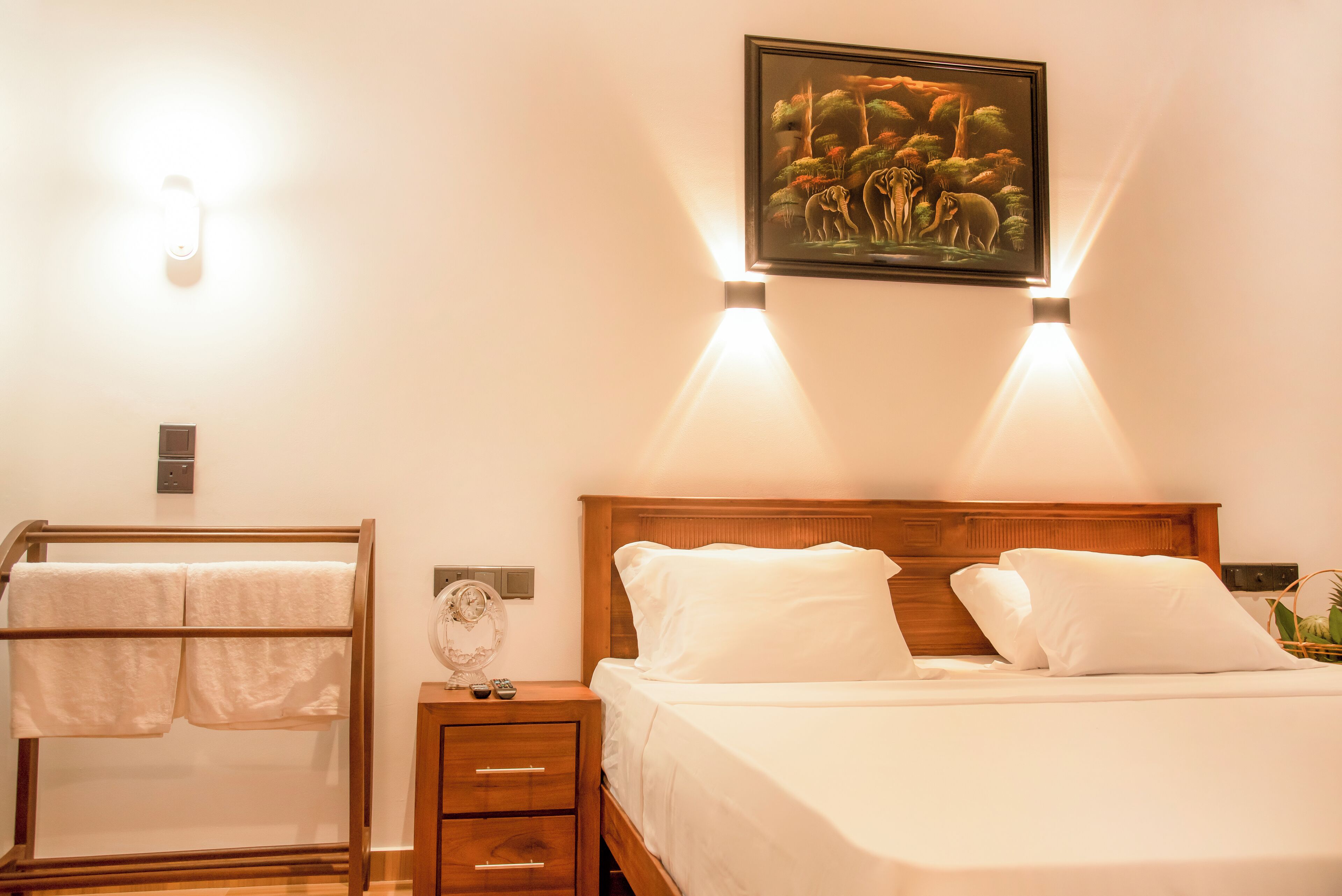 A hotel room with a bed, a bedside table with a lamp, a wall-mounted picture, and a towel rack.
