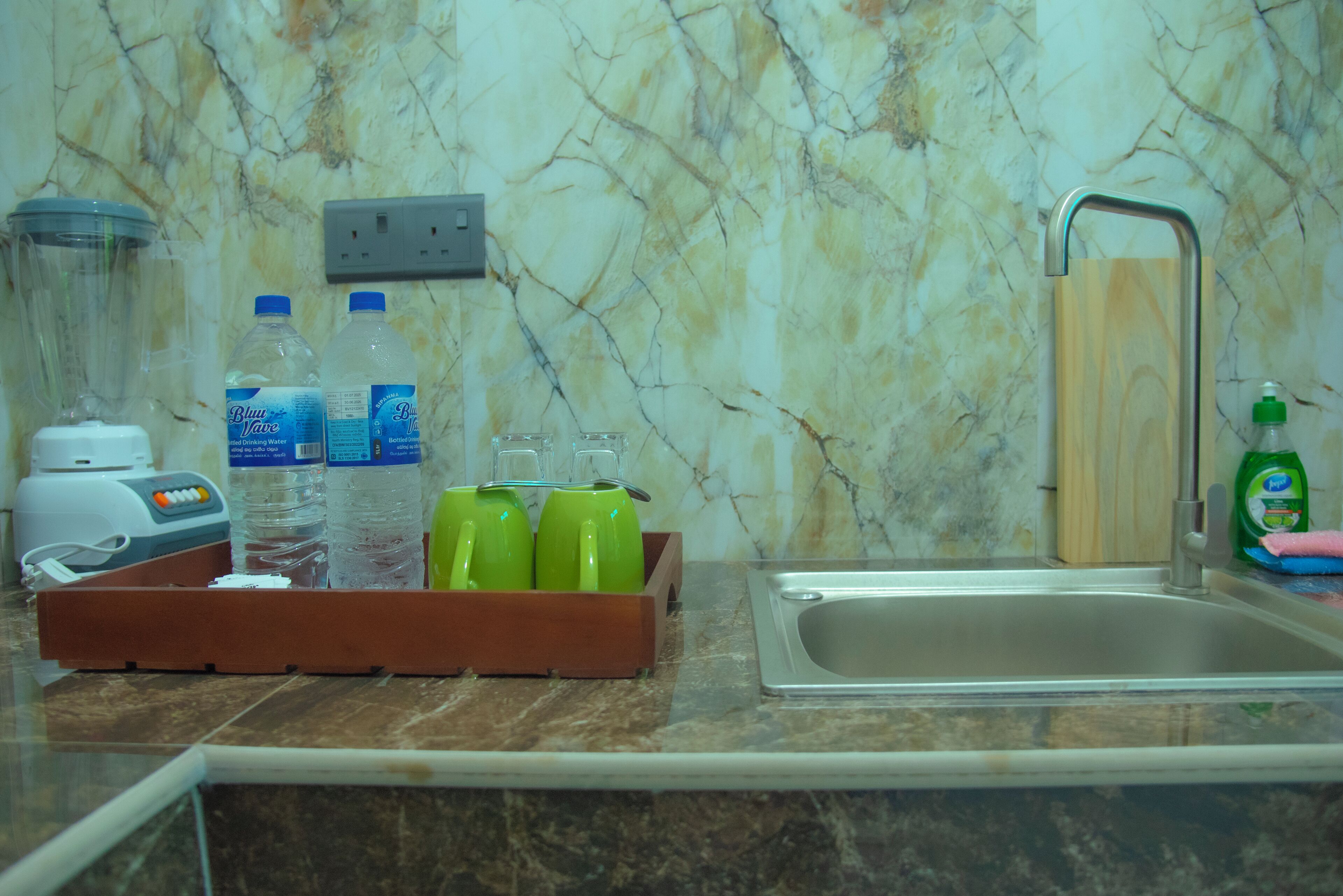 A kitchen counter with a marble backsplash, a sink, and a wooden tray holding water bottles and glasses.
