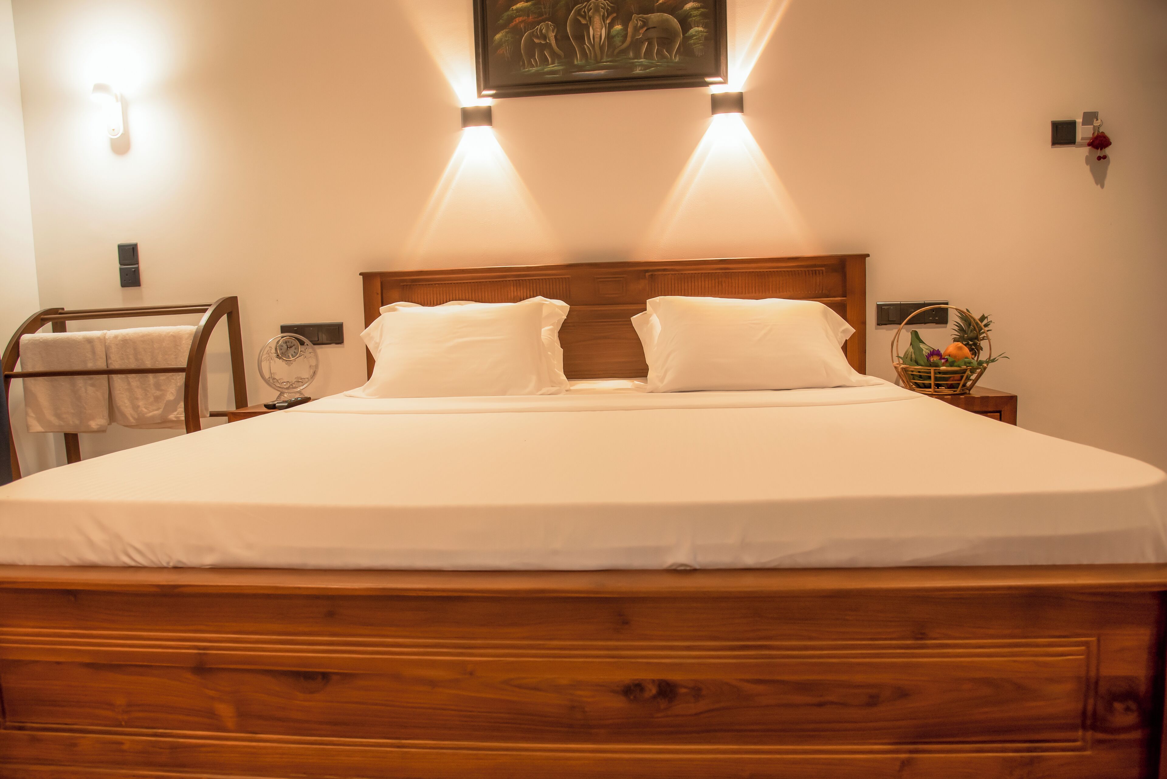 Un lit bien fait, avec une tête de lit en bois, deux tables de chevet avec des lampes, une œuvre d’art accrochée au mur et un panier de fruits posé sur la table de chevet.