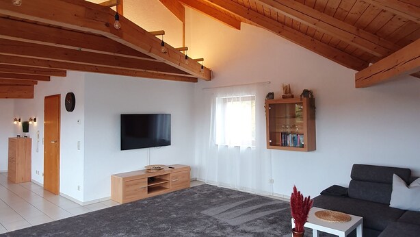 Un salón espacioso con techo alto, vigas de madera, alfombra gris, un sofá, una butaca, una mesita, un televisor y un mueble.