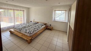 Un dormitorio con una cama de madera, una mesita de noche pequeña, una mesita auxiliar, una ventana y una puerta.