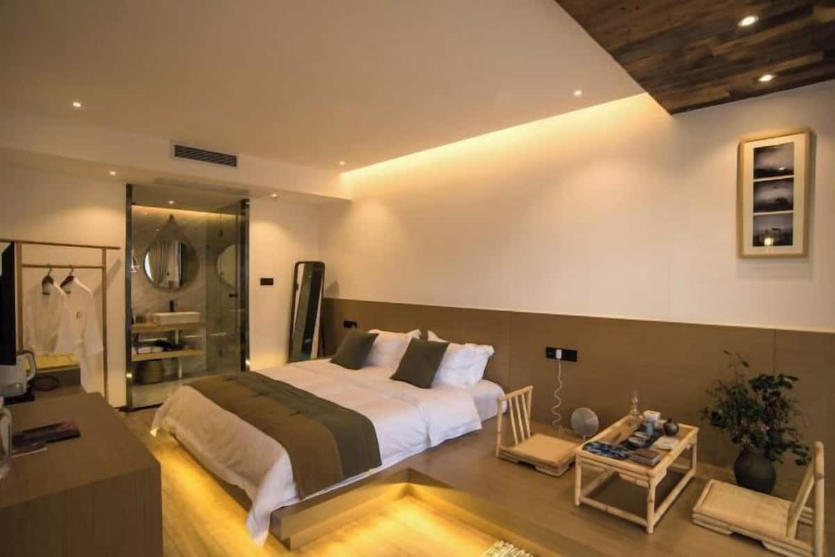 A modern hotel room with a large bed, a bedside table, a chair, a small table with a plant, and a mirror.