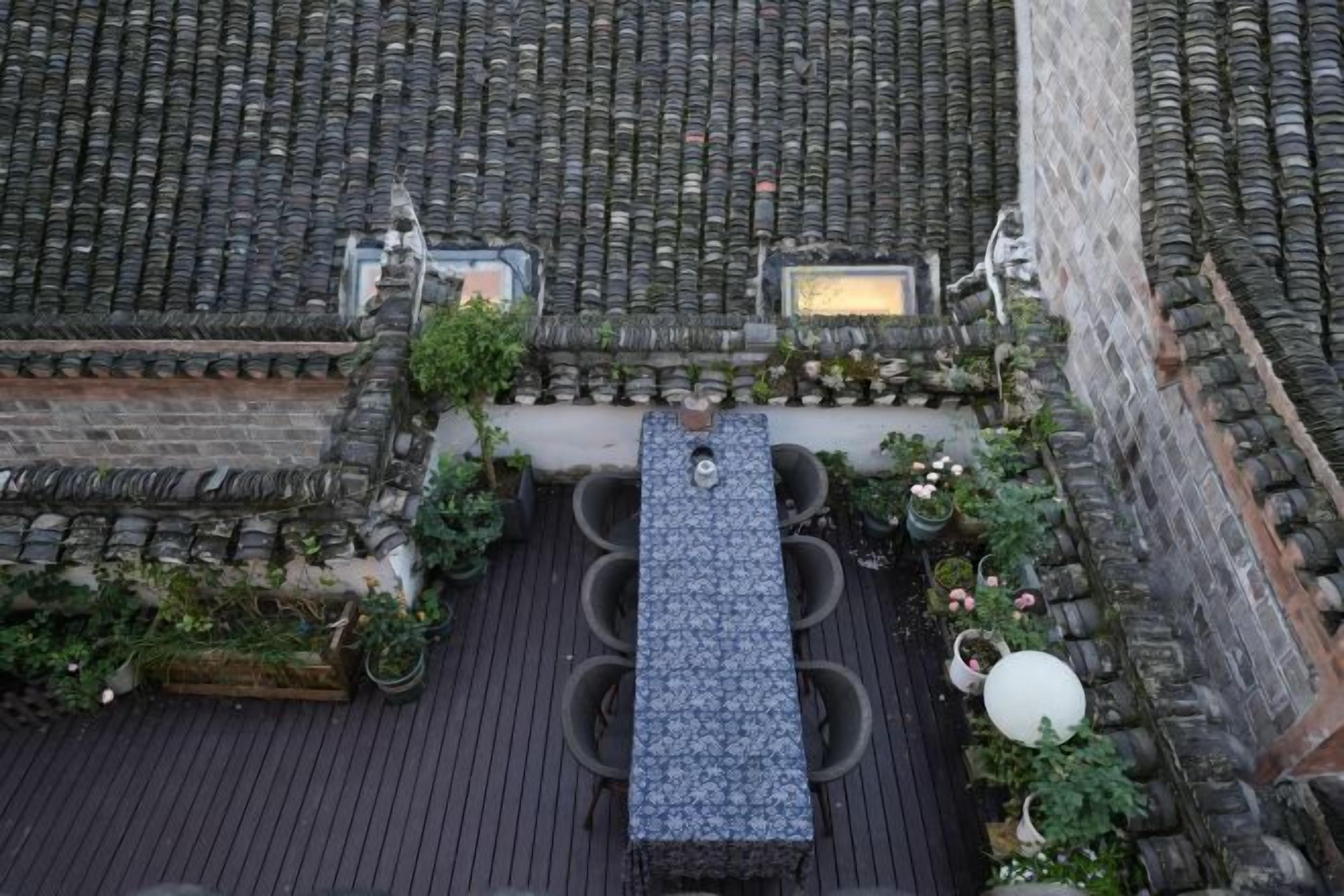 A rooftop terrace with a long table and chairs, surrounded by potted plants and a stone wall.
