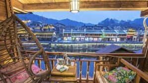 A wooden balcony with a wicker chair, a table with a kettle, and a view of traditional buildings across a river. - Feng Mian Su She Guesthouse (Fenghuang)