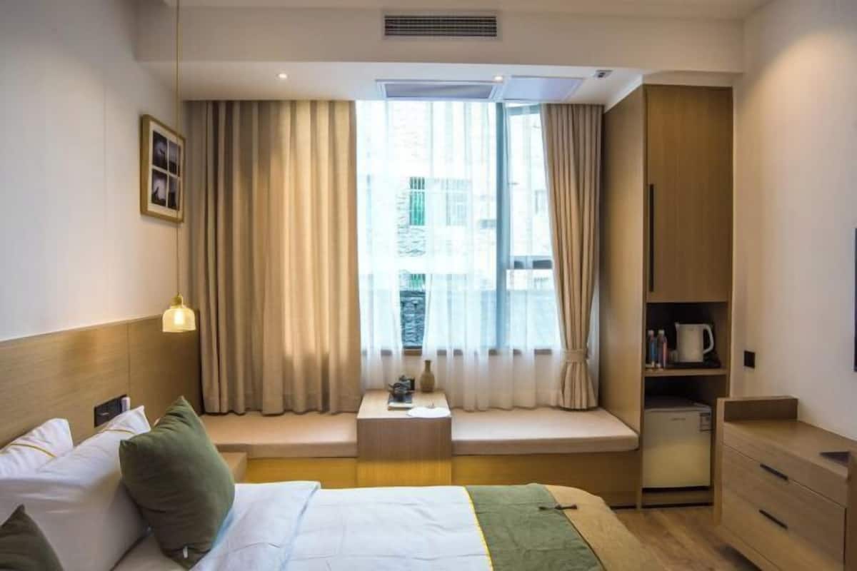 A hotel room with a bed, a small table with a lamp, a window seat, a wardrobe, and a mini-fridge.