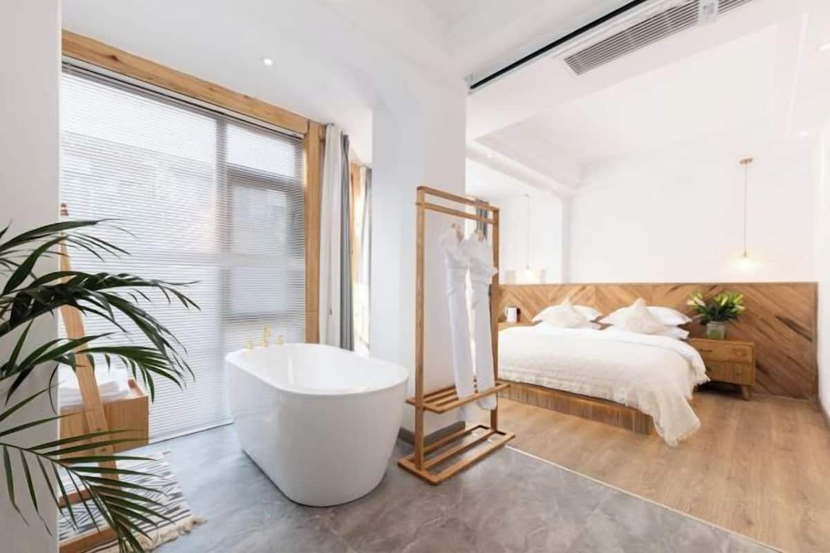 A modern bedroom with a large bathtub, a wooden headboard, and a large window with blinds.