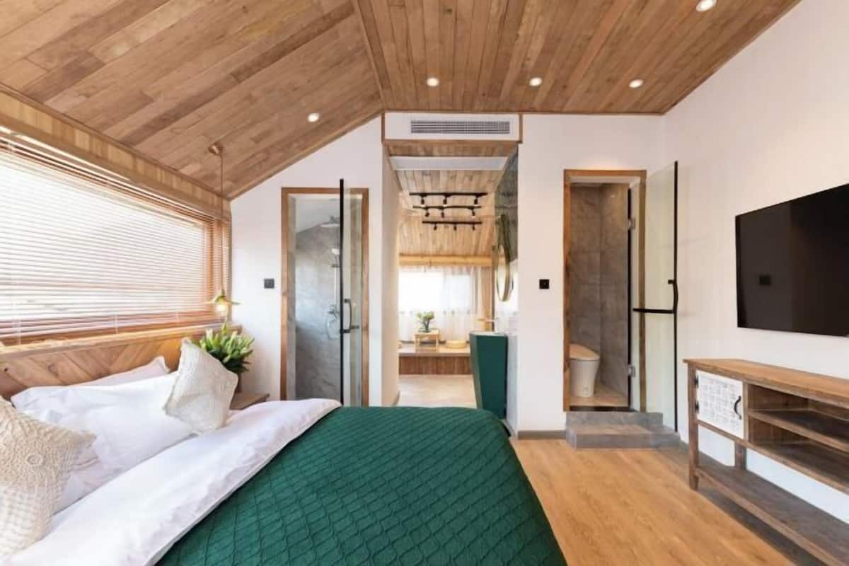A modern bedroom with a wooden ceiling, a large bed with green bedding, a TV on the wall, and a bathroom visible through an open door.