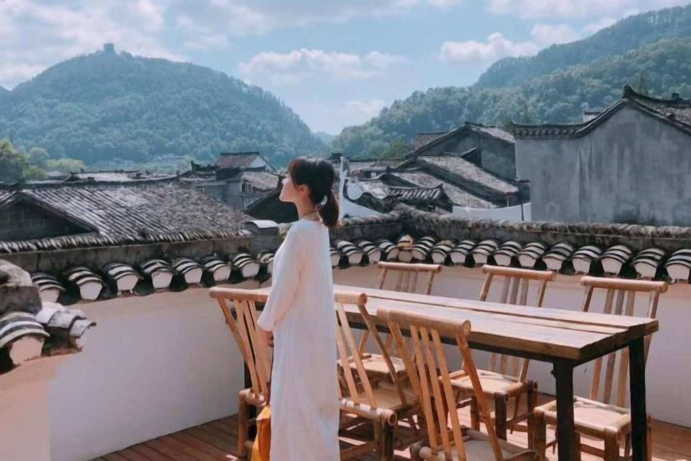 Une personne se tient sur une terrasse d’où l’on peut admirer des bâtiments traditionnels et un paysage montagneux.