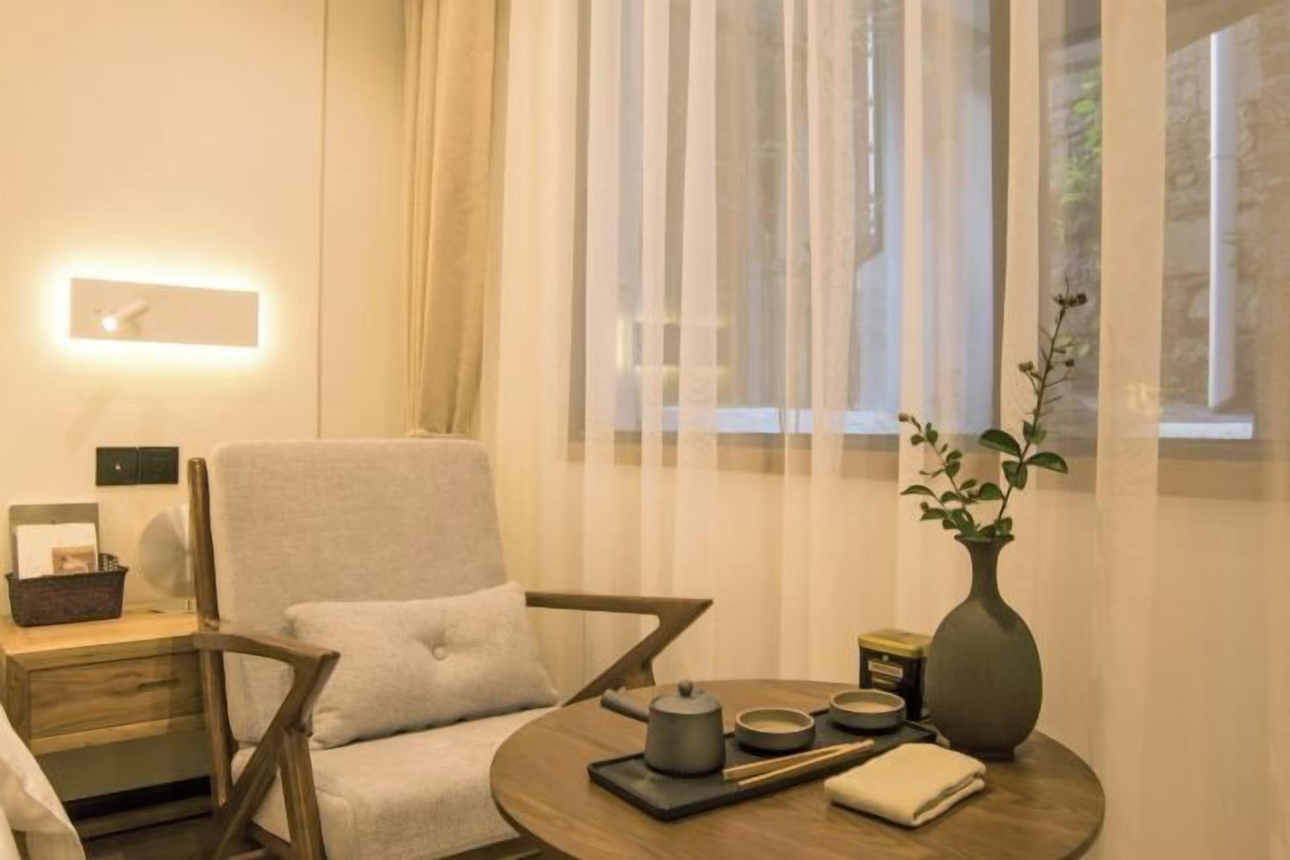A modern hotel room with a round wooden table, a comfortable armchair, a vase with a plant, and a bedside table with a lamp.