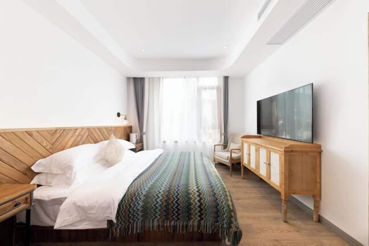 A hotel room with a wooden headboard bed, a large flat-screen TV, a wooden cabinet, and a view of the outdoors through the window.