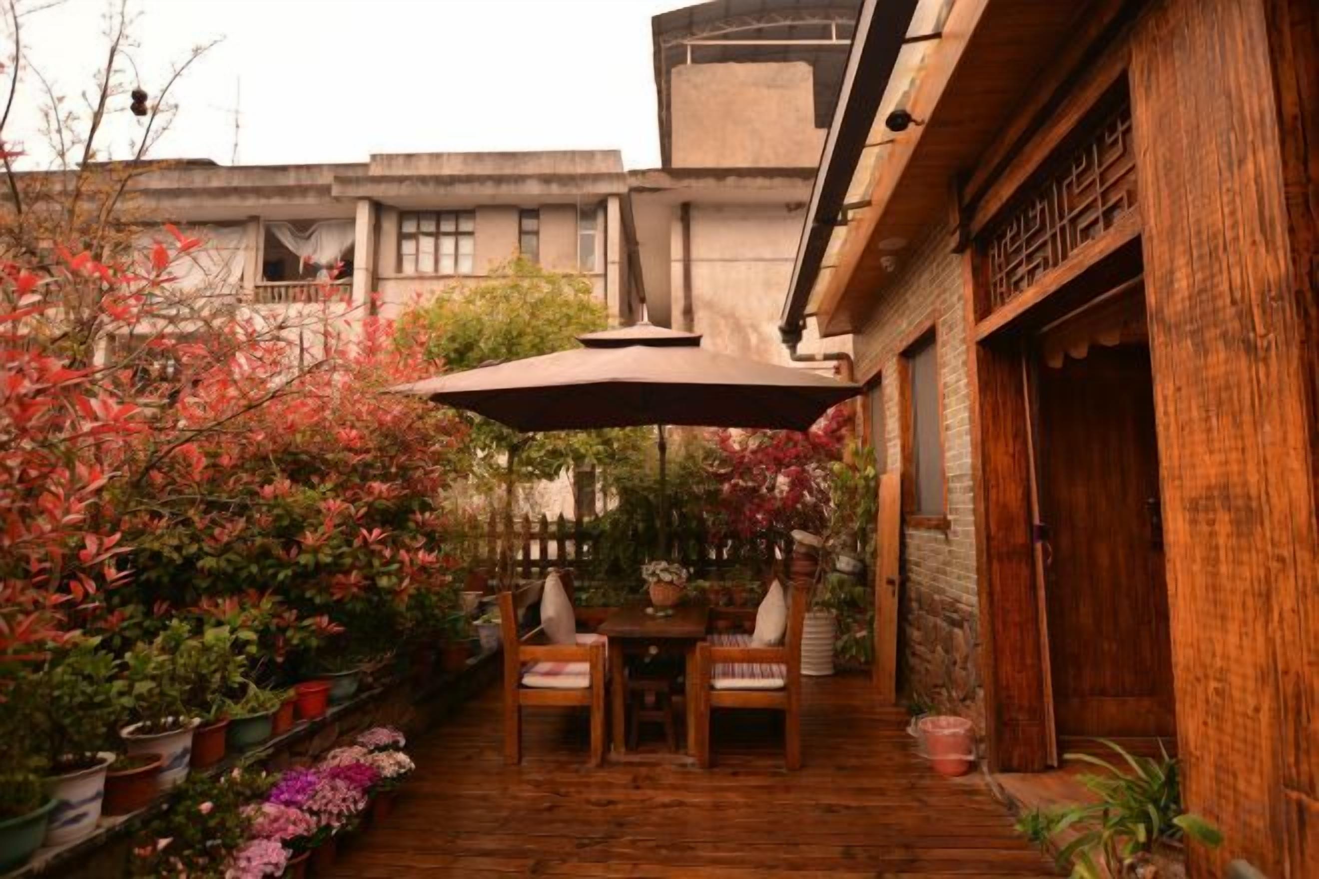 Une terrasse extérieure avec une table et des chaises en bois, entourée de plantes en pot et d’un grand parasol.