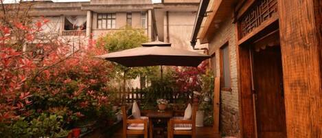Une terrasse extérieure avec une table et des chaises en bois, entourée de plantes en pot et d’un grand parasol.