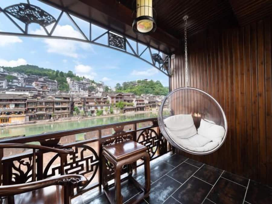 Un balcón de madera con una silla colgante, una mesa y vistas a un pueblo a orillas del río.